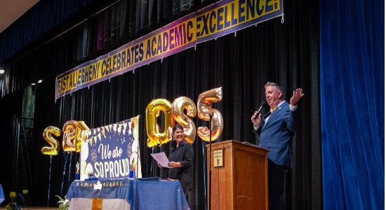  Principal stands at podium speaking into microphone to welcome crowd